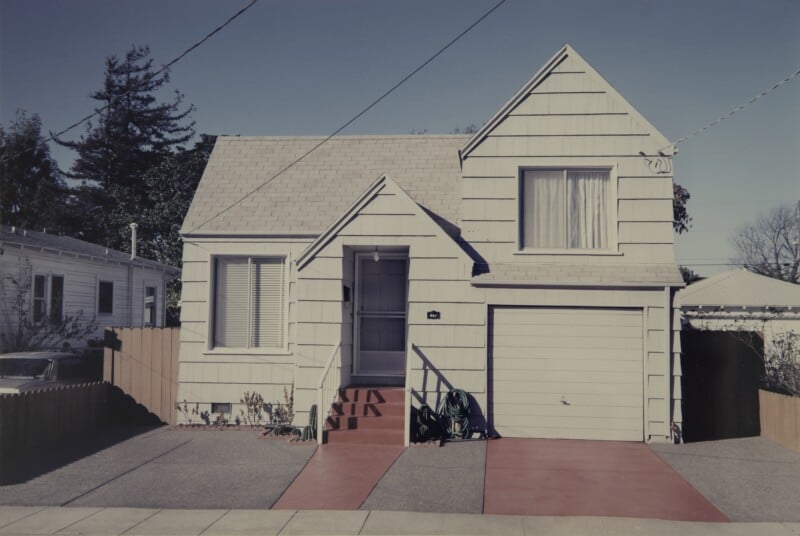A white, two-story house with a front-facing gable, a single-car garage, and a red concrete walkway and driveway apron.