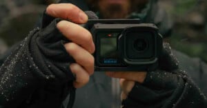 A person wearing black fingerless gloves holds a GoPro camera in a protective housing, with a small red light visible on the front.