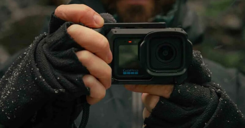 A person wearing black fingerless gloves holds a GoPro camera in a protective housing, with a small red light visible on the front.