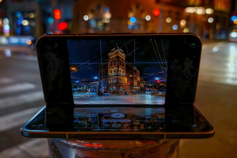 A foldable smartphone sits on a post at night, displaying a camera interface with a grid overlay and a long exposure setting while capturing a street scene with a historic building.