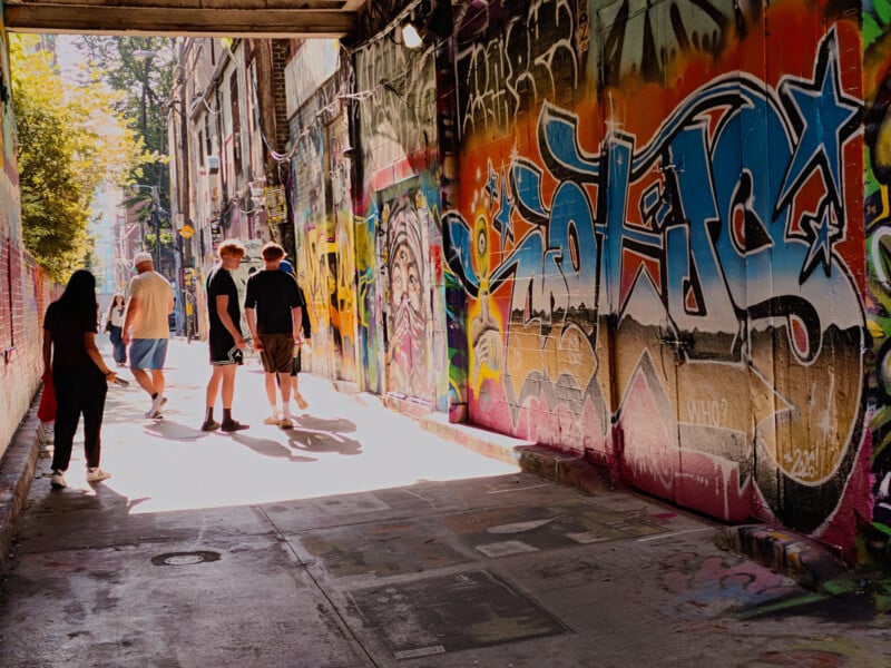 People walk through a narrow urban alleyway lined with colorful graffiti-covered brick walls.