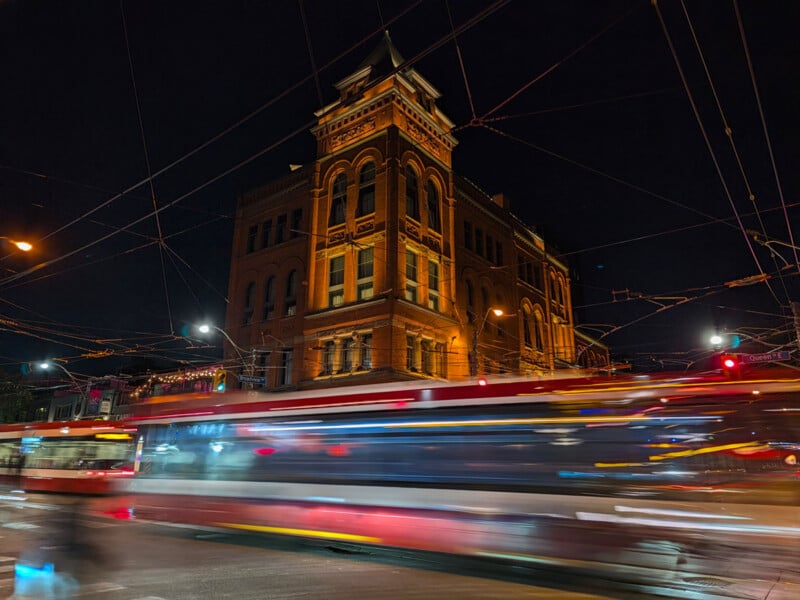 A streetcar moves rapidly through an intersection at night, creating a motion blur in front of a historic brick building with a corner tower.