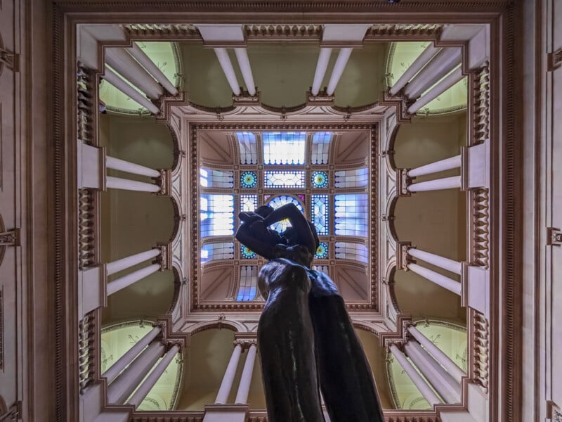 A low-angle view looking up at a bronze sculpture of two figures embracing, centered beneath a square, multi-paned stained glass skylight surrounded by classical columns and architectural molding.