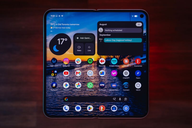 A foldable smartphone screen displays a home screen with a weather widget, a calendar widget, a Google search bar, and a grid of various app icons over a sunset harbor background.