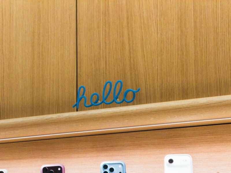 A blue, cursive "hello" sign is mounted on a light wood wall above a shelf displaying several smartphones.
