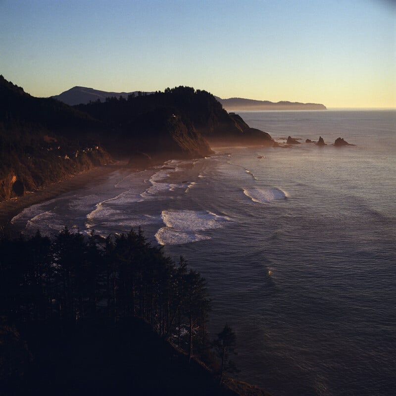 A high-angle view of a rugged coastline at sunset, with dark forested cliffs overlooking a beach and the ocean.