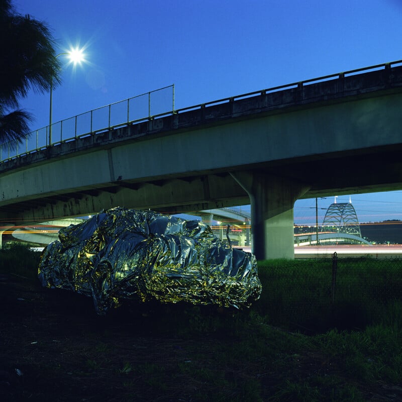 A car covered in a crinkled, reflective silver emergency blanket sits under a highway overpass at dusk, with a bridge visible in the distance.