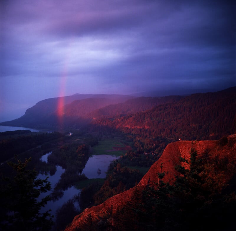 A vibrant sunset illuminates a cliffside and a distant rainbow over a river valley under a dark, cloudy sky.