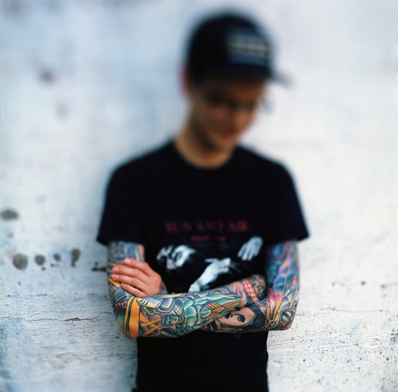 A person with heavily tattooed arms stands with their arms crossed, wearing a black t-shirt and a baseball cap.