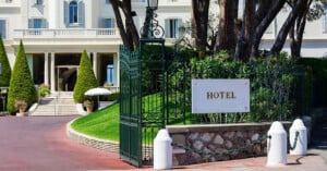 A white hotel building with a grand staircase and manicured cone-shaped trees sits behind a green wrought-iron gate with a sign that reads HOTEL.