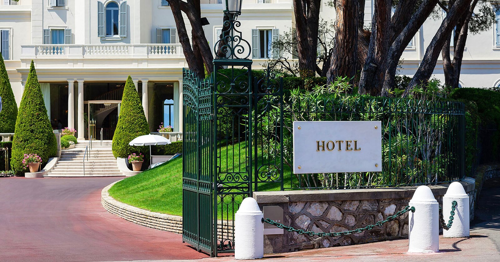 A white hotel building with a grand staircase and manicured cone-shaped trees sits behind a green wrought-iron gate with a sign that reads HOTEL.