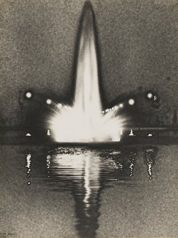 A black-and-white photograph shows a brightly illuminated fountain shooting a tall jet of water into the night air, with its reflection shimmering on the dark water below.