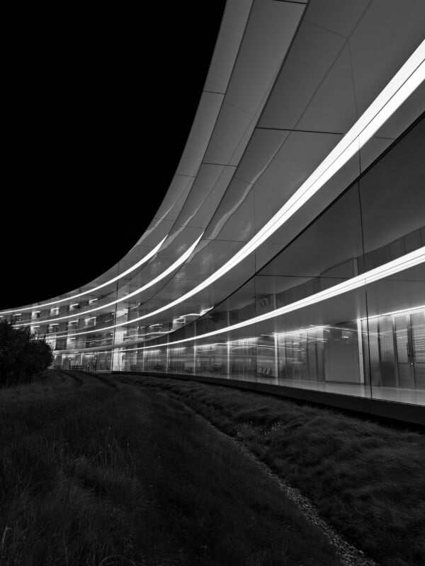 A black and white low-angle shot of a curved, modern glass building at night, featuring horizontal light strips and a grassy foreground.