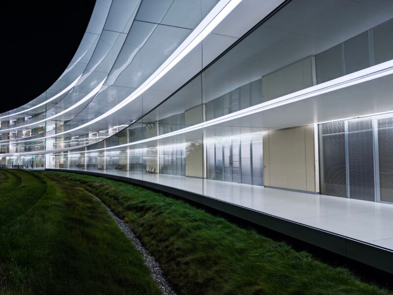 A curved, modern building with floor-to-ceiling glass walls and illuminated horizontal light strips overlooks a grassy embankment at night.