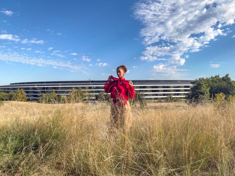 A person with curly hair wearing a red blouse and tan pants stands in a field of tall, dry grass in front of a long, modern building under a blue sky with scattered clouds.