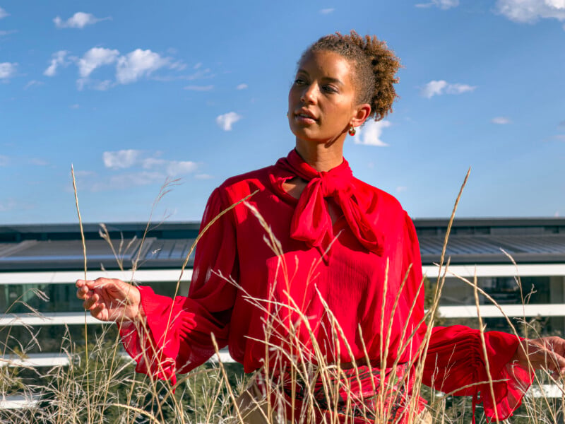 A woman with curly hair wears a long-sleeved red blouse with a bow at the neck while standing in a field of tall grass against a blue sky and a modern building.