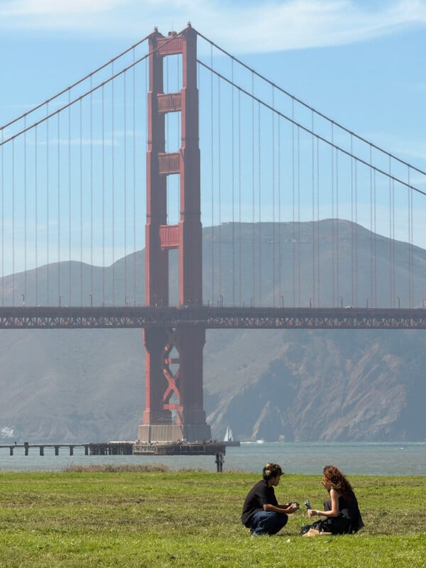 Two people sit on a grassy lawn in front of the Golden Gate Bridge under a clear blue sky.