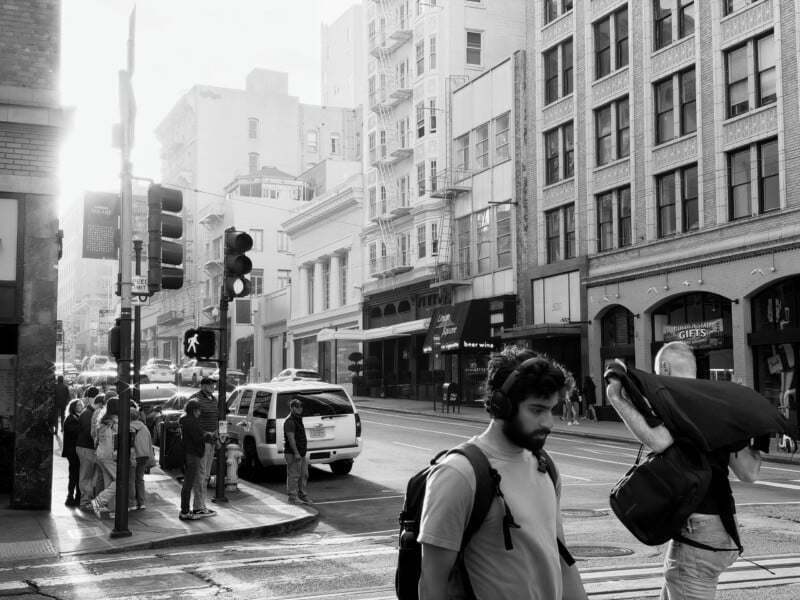 Pedestrians wait at a street corner in a city while a man with a backpack and headphones walks across the foreground.