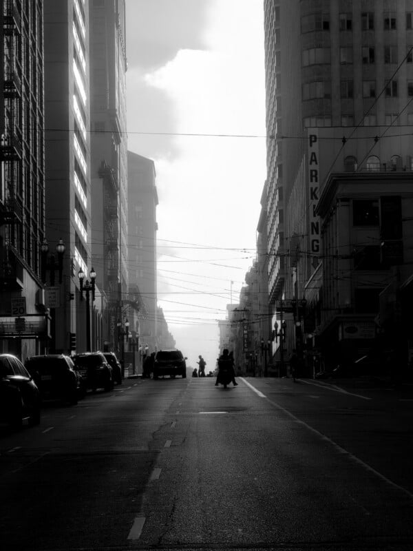 A black and white street-level view looking down a city street lined with tall buildings, with a motorcycle and cars driving toward a bright, hazy sky.