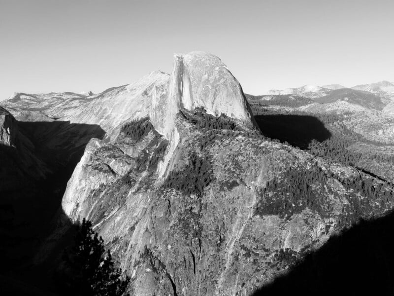 A black and white landscape view of the sheer granite face of Half Dome rising above a forested mountain ridge.