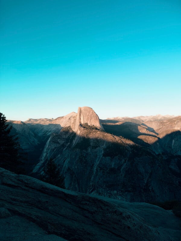 A granite mountain peak with a sheer vertical face rises above a deep, shadowed valley under a clear blue sky.