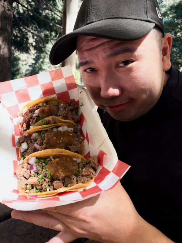 A man wearing a black baseball cap holds a red and white checkered paper tray containing three street tacos topped with meat, onions, cilantro, and salsa.