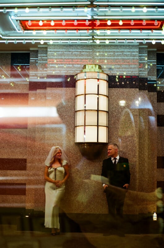 A woman in a white dress and veil stands next to a man in a black suit in front of an Art Deco wall sconce and a marquee with light bulbs.