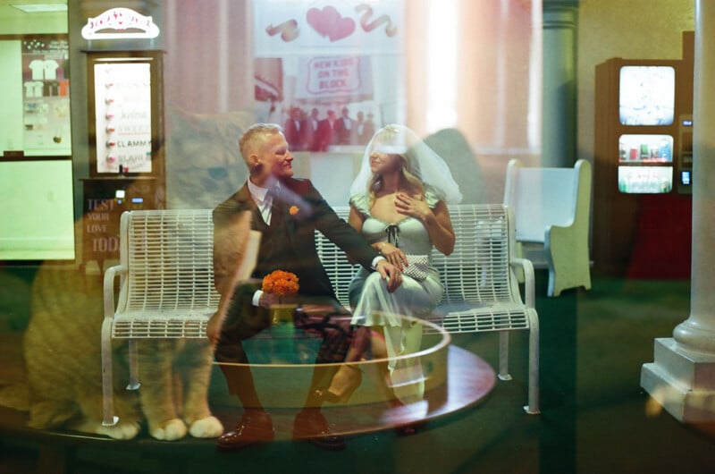 A bride and groom sit on a white metal bench, viewed through a glass reflection that shows a faint, large cat-like shape in the foreground.