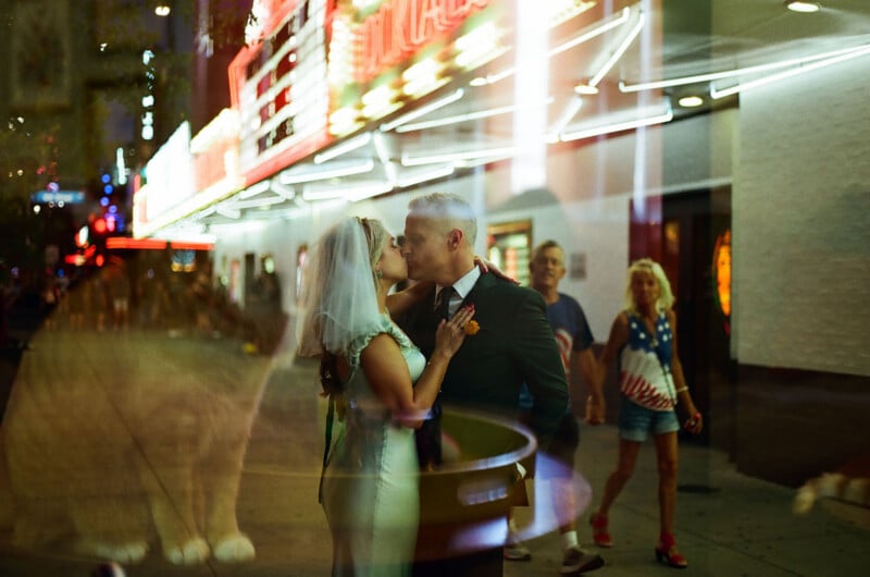 A bride and groom kiss in front of a brightly lit theater marquee, viewed through a window with reflections of people walking by.