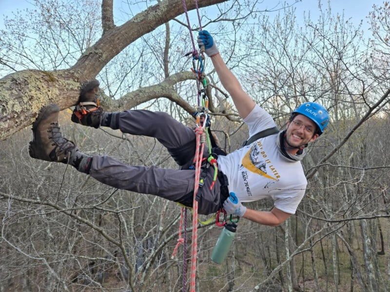 A smiling man wearing a blue helmet and climbing gear is suspended in a tree, holding onto a rope system with one hand.