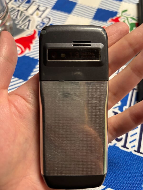 A hand holds a rectangular, silver and black mobile phone with a 1.3-megapixel camera lens visible on the back.