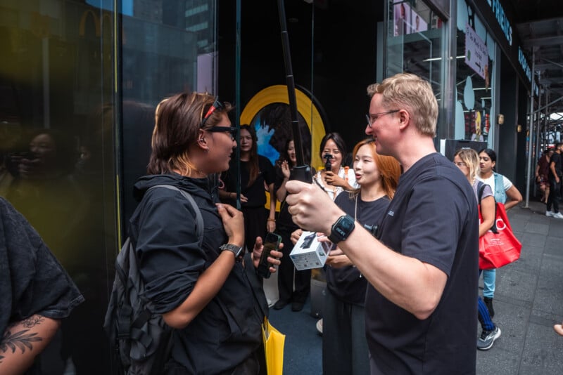 A man holding a selfie stick and a product box speaks to a person holding a smartphone outside a storefront, with several other people standing nearby.