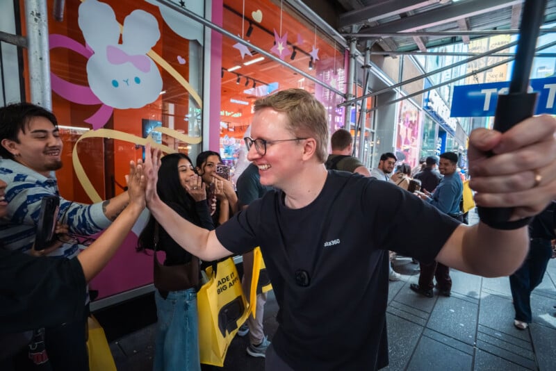 A man in a black t-shirt high-fives a person while holding a camera on a pole, with onlookers and a storefront featuring a cartoon rabbit graphic in the background.