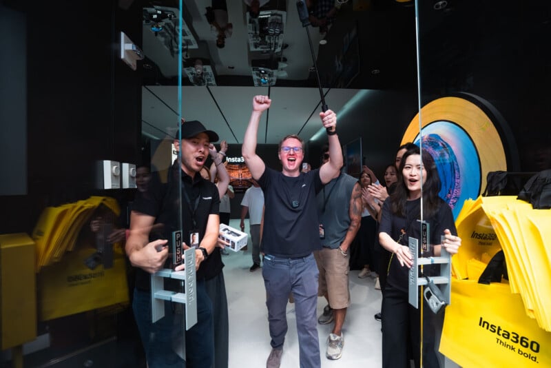 A group of people cheer as they walk through glass doors into an Insta360 store, with one man in the center holding a camera on a selfie stick.