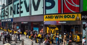 A crowd of people stands on a sidewalk in front of storefronts for Tissot and Insta360, with a large "AND WIN GOLD" sign above.