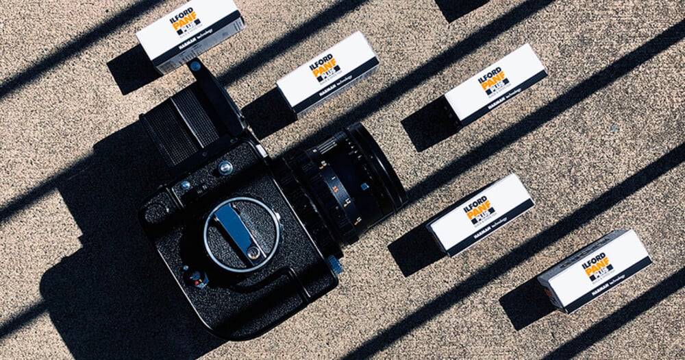 A medium format camera sits on a concrete surface surrounded by four boxes of Ilford Pan F Plus black and white film, all cast in long, diagonal shadows.