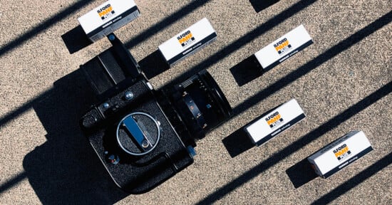 A medium format camera sits on a concrete surface surrounded by four boxes of Ilford Pan F Plus black and white film, all cast in long, diagonal shadows.