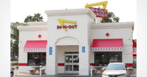 An In-N-Out Burger restaurant building with a white facade, red awnings, and a yellow arrow sign, featuring outdoor picnic tables and a parked car.