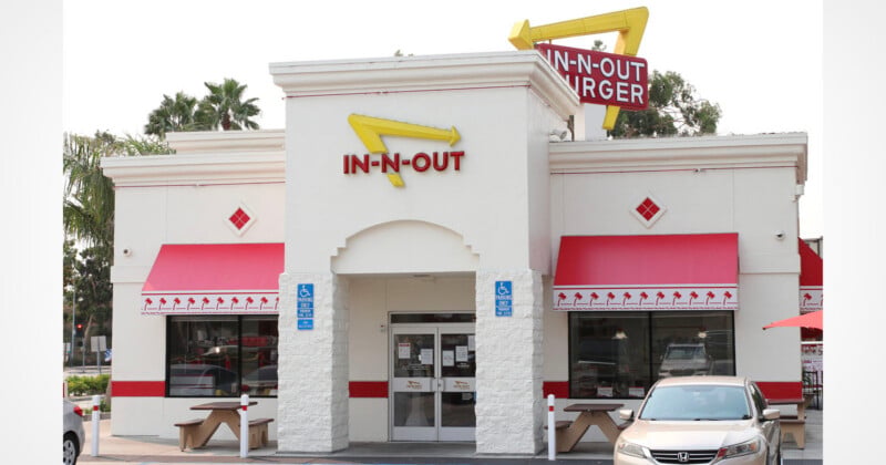 An In-N-Out Burger restaurant building with a white facade, red awnings, and a yellow arrow sign, featuring outdoor picnic tables and a parked car.
