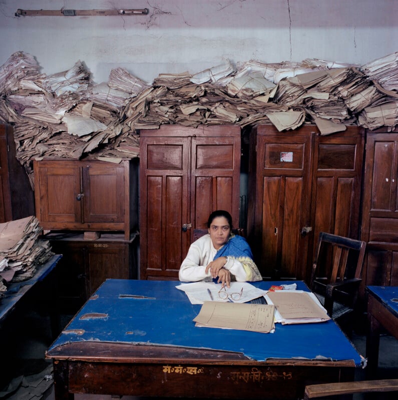 A woman sits at a desk covered in documents, surrounded by tall wooden cabinets stacked high with piles of paper files.