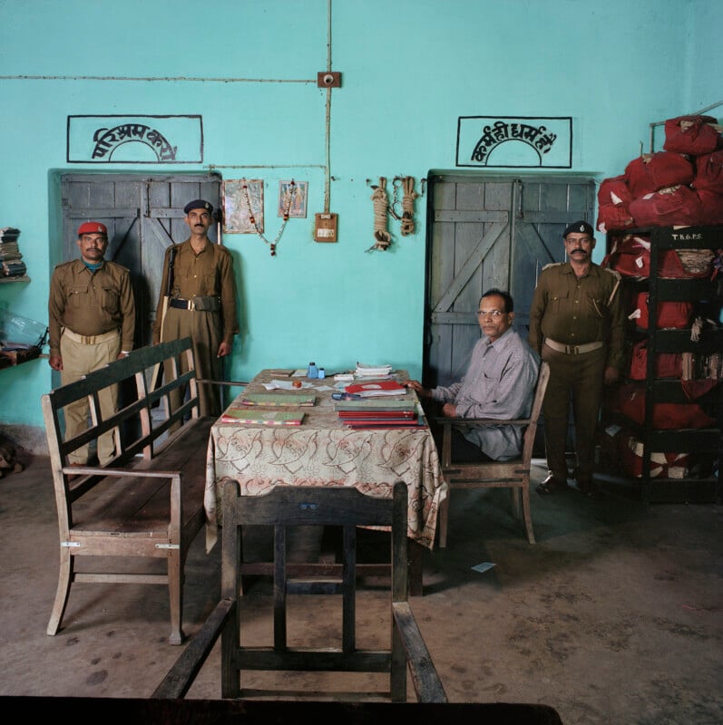 Three uniformed police officers stand in an office with a man seated at a desk covered in paperwork and files.