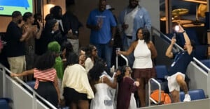 A group of people in a stadium suite celebrate, with one man standing on a chair to take a photo of a woman in a white halter top.
