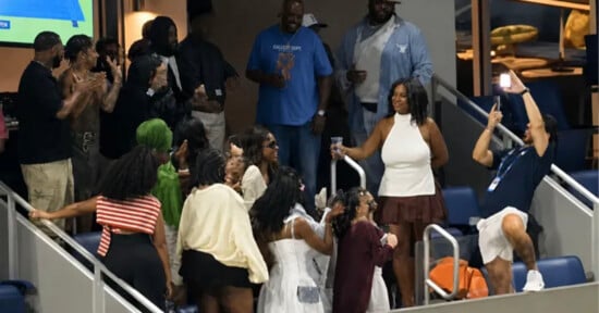 A group of people in a stadium suite celebrate, with one man standing on a chair to take a photo of a woman in a white halter top.