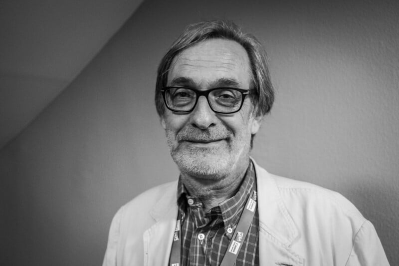 A middle-aged man with graying hair, a beard, and glasses smiles at the camera while wearing a plaid shirt and a light-colored jacket.