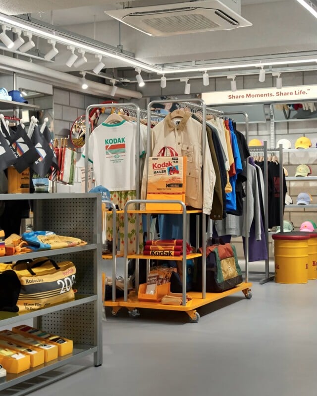 A retail store interior features racks of Kodak-branded clothing, bags, and accessories, with yellow display carts and shelves against a grey brick wall.