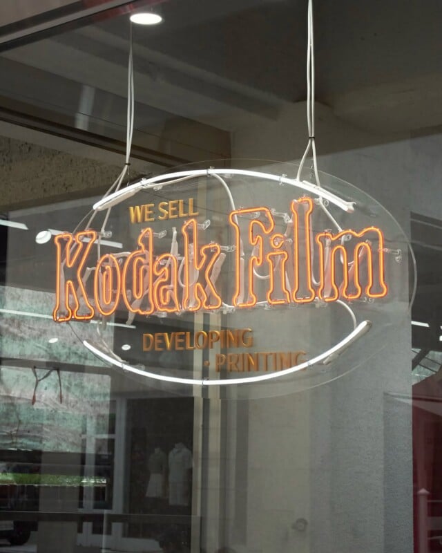 A neon sign hanging in a window reads "WE SELL Kodak Film" in orange, with "DEVELOPING" and "PRINTING" printed below in gold.