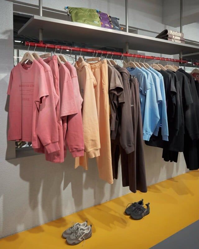 A clothing rack displays shirts, hoodies, and pants in a gradient of colors from pink to black, with two pairs of sneakers on the yellow floor below.