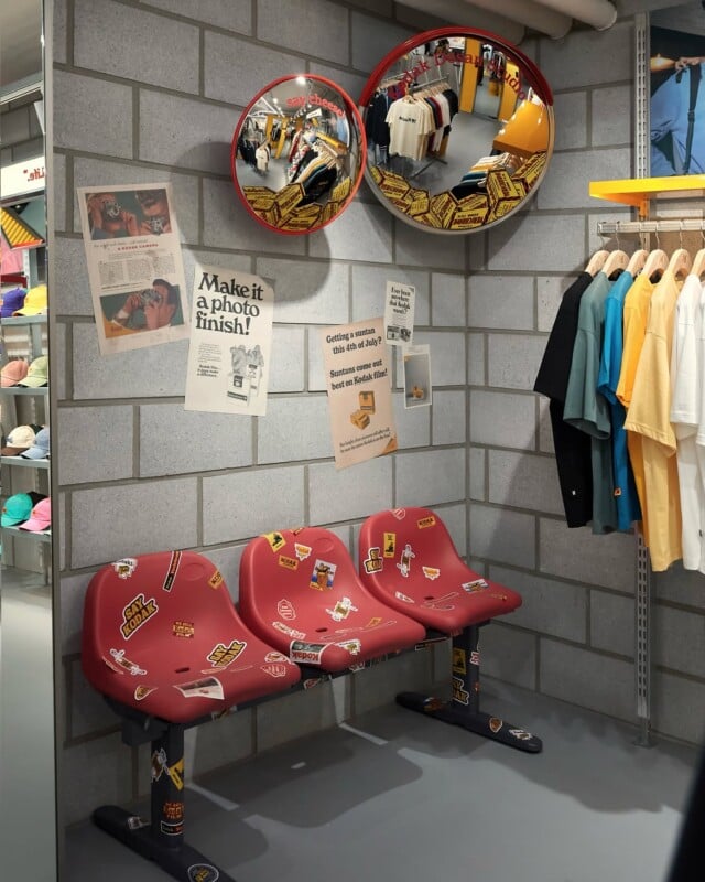 A row of three red plastic chairs decorated with stickers sits against a gray brick wall in a retail store, with vintage-style posters and two circular convex mirrors mounted above.