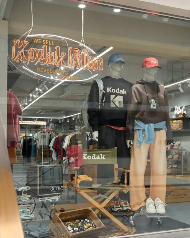 A storefront window displays two mannequins wearing Kodak-branded clothing, with a neon sign above reading "We Sell Kodak Film.