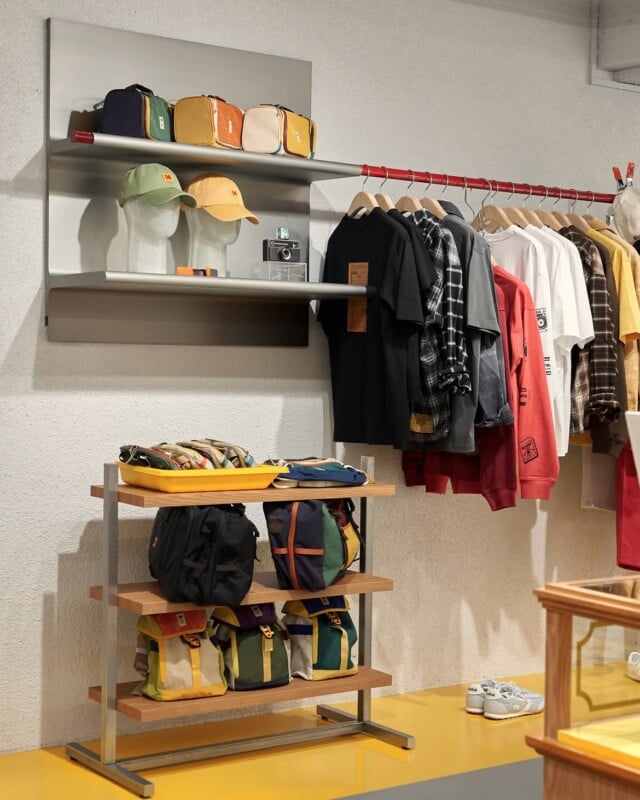 A retail display features shelves with bags, hats, and a camera, alongside a clothing rack with shirts and jackets, and a wooden shelving unit holding various backpacks.